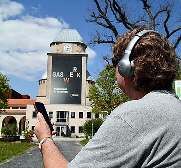 Person mit Kopfhörern auf dem Kopf und einem Smartphone in der Hand steht vor dem Gaswerk.