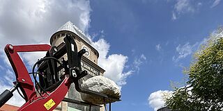 Ein massiver Stein wird von einem Gabelstapler auf Eisenstäbe gehoben. Im Hintergrund ist das Ofenhaus und der Gaskessel auf dem historischen Gaswerk in Augsburg zu erkennen.