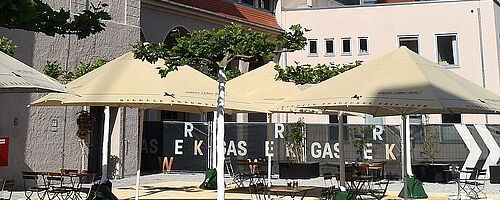 Gaswerksareal mit Sitzmöglichkeiten, Tischen und Sonnenschirmen. Weiterverlinkung auf Gaswerksareal Newsartikel