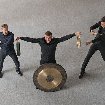 Drei Männer vor einem großen Gong - Cover von Schlag3