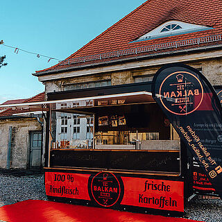 Frontansicht auf Balkan Kitchen mit Bannern und roten Teppich auf dem Boden. Weiterverlinkung auf Magazin Artikel zu Balkan Kitchen