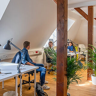 Gaswork Coworkingspace. Sechs Menschen sitzen in einem Büro, teilweise an einem Schreibtisch mit einem Laptop. Im Büro sind Pflanzen. Weiterverlinkung auf Gaswerk kreative Arbeit Seite
