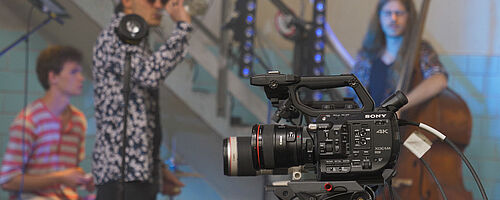 Filmkamera. Im Hintergrund sind Leute mit Instrumenten. Weiterverlinkung auf Stay Home Festival Newsartikel