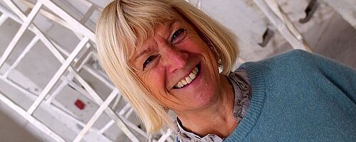 Lachende Frau steht in einem Gebäude vor Skulpturen. Weiterverlinkung auf INTERFERENZEN Newsartikel