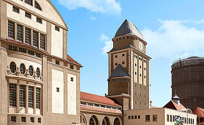 Gaswerkgelände an einem sonnigen Tag mit Besuchern. Weiterverlinkung auf Staatstheater Newsartikel