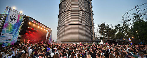 Das Bild zeigt eine feiernde Menschenmenge vor einer großen Bühne. Die Bühne ist mit Scheinwerfern rot beleuchtet. Links neben der Bühne ist ein Banner zu sehen, auf dem Modular Festival steht. Auf dem Banner kann man lesen, dass das Festival von Sponsoren wie den Stadtwerken Augsburg (swa) gefördert wird. Im Hintergrund des Bilds ist der Gaskessel zu sehen.