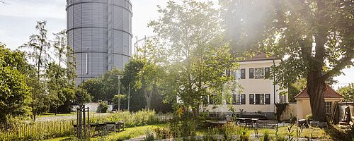 Gaskessel im Gaswerk. Im Vordergrund sind Bäume und ein Gebäude.