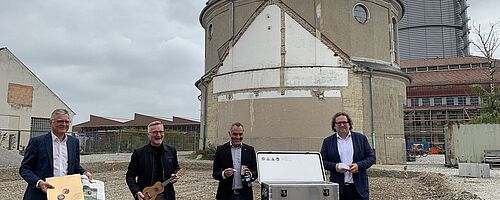 Vier Männer stehen mit Zeitungen und Instrumenten vor einem Tisch auf dem eine Box steht. Weiterverlinkung auf Musikbox Newsartikel