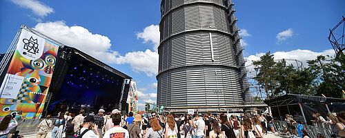 Menschen stehen auf dem Gaswerksgelände. Im Hintergrund ist eine Bühne und der große Gasbehälter. Weiterverlinkung auf swa und Modular Partnerschaft Newsartikel