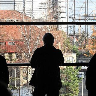 Menschen stehen vor einem Fenster mit Blick auf den Gaskessel auf dem Gaswerksgelände. Weiterverlinkung auf die Gaswerk Führungen Seite