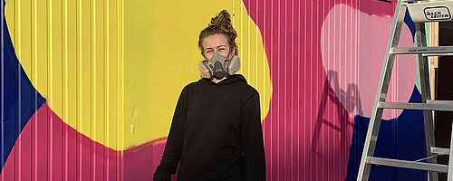 Frau mit Atemschutzmaske und Spraydose steht vor einem bemalten WC Container. Weiterverlinkung auf WC Container Newsartikel