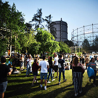Menschen laufen auf der Wiese des Gaswerksgelände. Im Hintergrund ist der Gaskessel. Weirerverlinkung auf die Veranstaltungskalender Seite