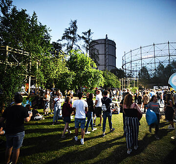 Menschen laufen auf der Wiese des Gaswerksgelände. Im Hintergrund ist der Gaskessel. 