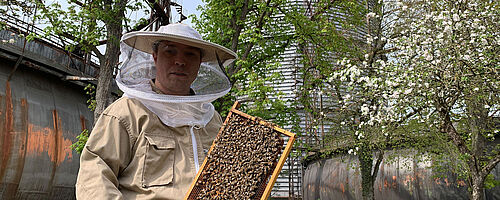 Imker mit Bienenwabe vor dem Gaswerk. Weiterverlinkung auf neue Bewohner fürs Gaswerksgelände Newsartikel