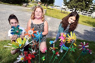 Drei Mädchen stecken gebastelte Blumen in eine Wiese auf dem Gaswerksgelände. - Beim anklicken wird das Bild vergrößert.