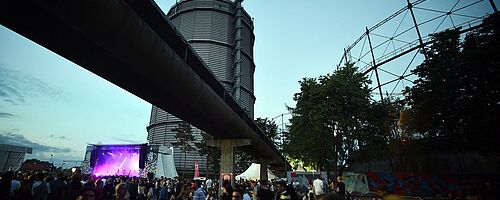 Menschen laufen auf dem Gaswerk im Hintergrund ist eine beleuchtete Konzertbühne und der Gaskessel. Weiterverlinkung auf Festivalsommer Newsartikel