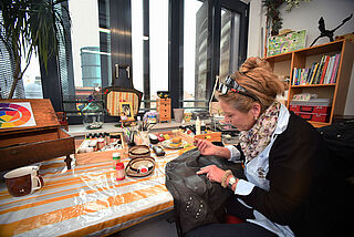 Frau sitzt an einem Tisch mit verschiedenen Farbutensilien und arbeitet an einer Lederjacke.