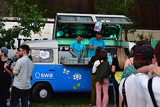 Personen stehen vor dem swa Wasser Bulli Schlange. Zwei Personen mit blauen Tshirts verteilen aus dem Bulli Wasser