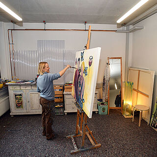 Frau steht in einem Raum mit Pinsel in der Hand vor einer Leinwand und malt ein Gemälde. Weiterverlinkung auf Gaswerk Raumanfrage