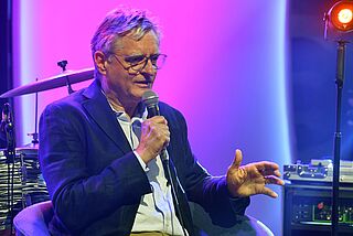 Mann mit Mikrofon sitzt vor Instrumenten. 