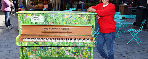 Bemaltes Klavier, in Grün Tönen. Frau lehnt sich an das Klavier an. 