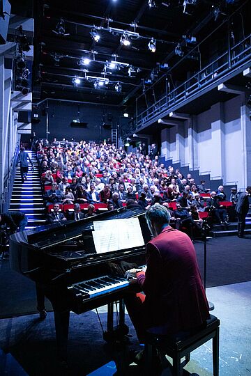 Pianist spielt auf einem Flügel mit Blick auf das volle Publikum der Brechtbühne