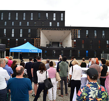 Menschen stehen an Stehtischen. Im Hintergrund ist eine Bühne in einem Gebäude und ein Stoffpavillon 