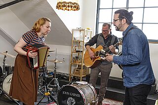Eine Frau und zwei Männer in einem Raum voller Musik-Equipment. Beide Männer spielen Gitarre und die Frau spielt Akkordeon. - Beim anklicken wird das Bild vergrößert. 