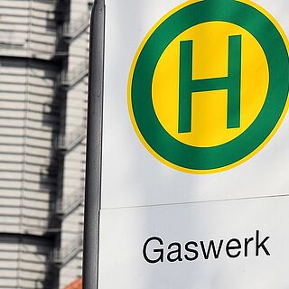 Gaswerk Haltestellenschild. Im Hintergrund ist ein Gebäude des Gaswerks. Weiterverlinkung auf Gaswerk Anfahrt Seite