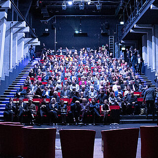 Mann sitzt auf einer Bühne an einem Klavier. Vor ihm sind aufsteigende Plätze auf denen Menschen sitzen. Weiterverlinkung auf Gaswerk Staatstheater Seite