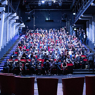 Mann sitzt auf einer Bühne an einem Klavier. Vor ihm sind aufsteigende Plätze auf denen Menschen sitzen. Weiterverlinkung auf Gaswerk Staatstheater Seite